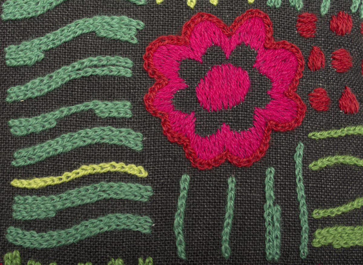 Stoppad kudde. Broderad på svart linne (Moralinne färg 2) med kedjesöm och plattsöm. Motivet består av blommor och ränder som gör att broderiet ser ut att vara indelad i rutor.
 Sytt i grönt, ljusgrönt, rött, cerice och orange. Baksida av samma tyg som framsidan.