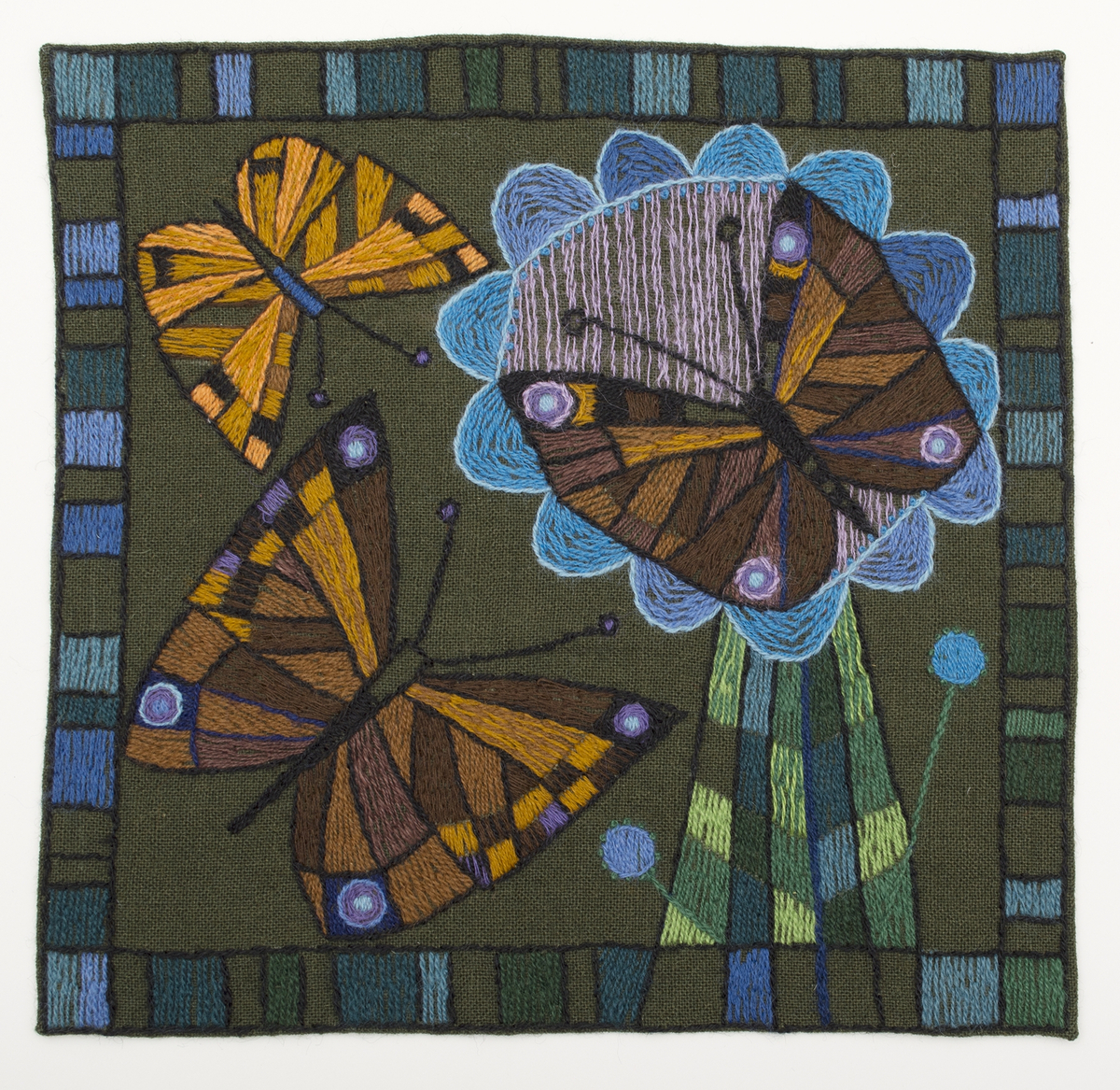 Fyrkantig bonad på brunt ylletyg, broderad med tvåtrådigt ullgarn i bruna nyanser, blålia nyanser och gröna nyanser. Fritt broderi med gles/tät schattersöm, stjälksöm, knutsöm. Motiv med tre fjärilar, varav en sitter på en blomma, ram i broderade rutor, omväxlande fyllda/ofyllda, 20 mm in från kanten. Redgarn, broderullgarn, Bergå ullgarn, lingarn 16/2.