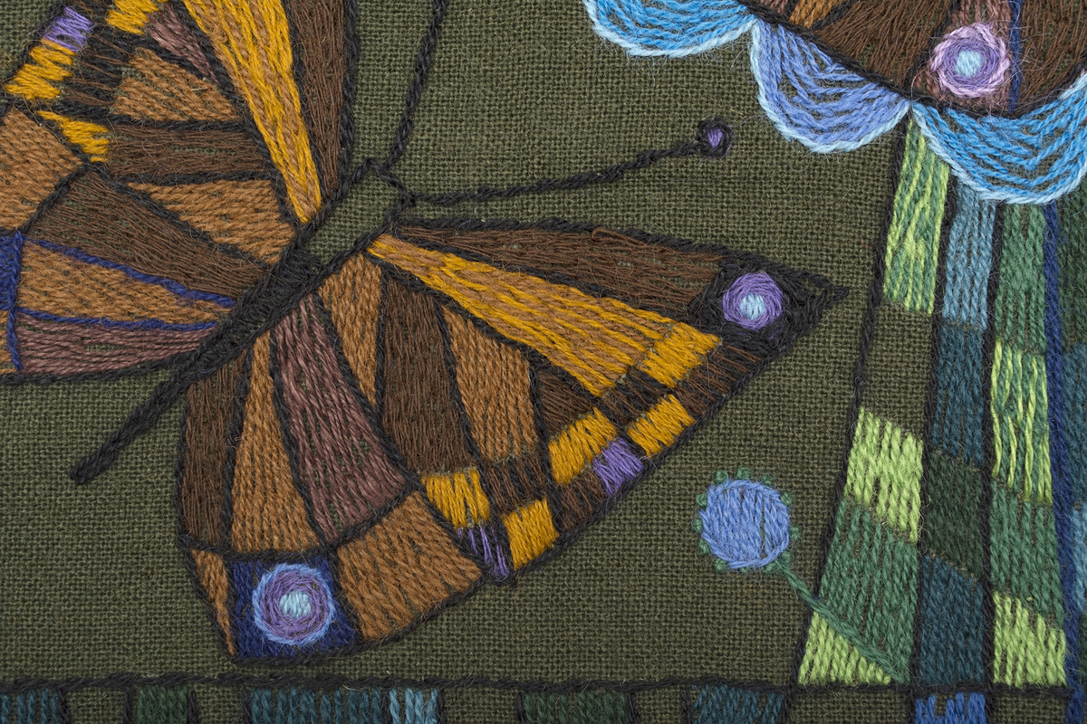 Fyrkantig bonad på brunt ylletyg, broderad med tvåtrådigt ullgarn i bruna nyanser, blålia nyanser och gröna nyanser. Fritt broderi med gles/tät schattersöm, stjälksöm, knutsöm. Motiv med tre fjärilar, varav en sitter på en blomma, ram i broderade rutor, omväxlande fyllda/ofyllda, 20 mm in från kanten. Redgarn, broderullgarn, Bergå ullgarn, lingarn 16/2.