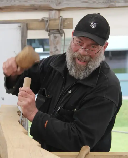 En mann med skjegg, briller, mørk jakke og caps holder en meisel og slår på den. 