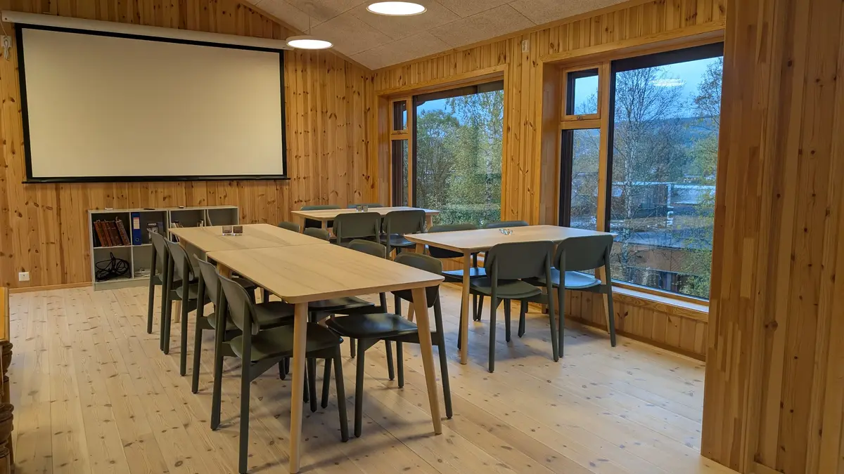 Bilde av kaferommet på Trysil-bygdetun. Det er satt opp et langbord på midten og bord inntil vinduene på høyre side. Lerret til prosjektoren er rullet ned. 