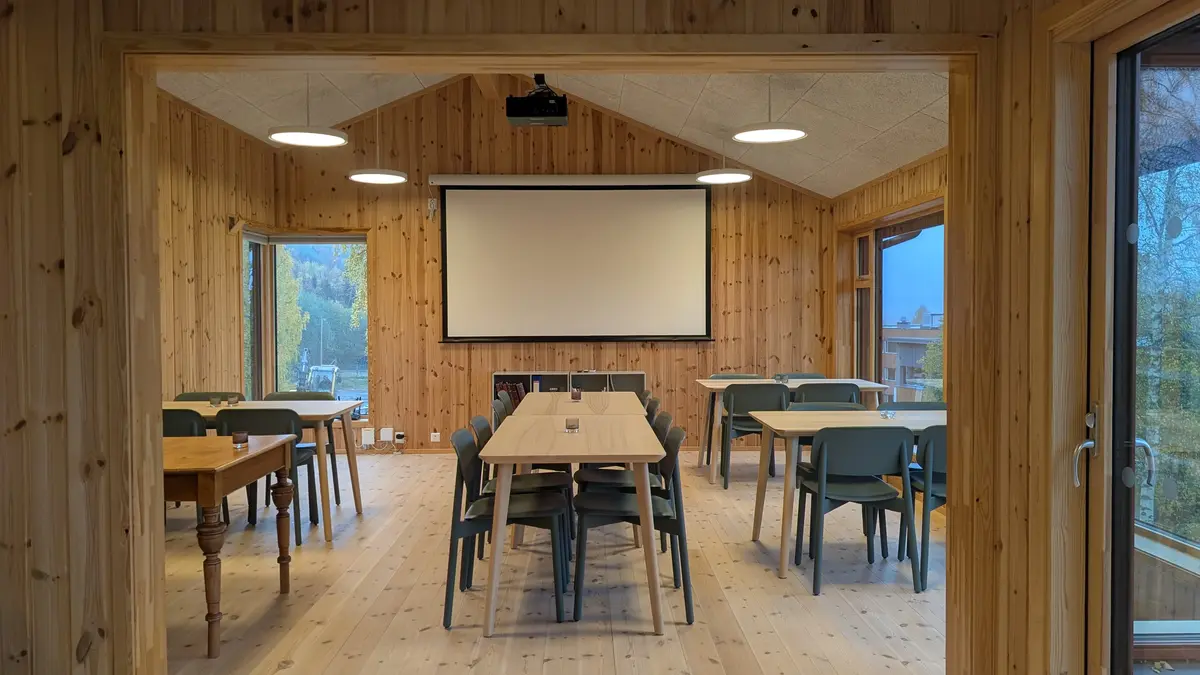 Bilde av kaferommet på Trysil-bygdetun. Det er satt opp et langbord på midten og på hver side står det enkelt bord inntil veggen og vinduer. Lerret til prosjektoren er rullet ned