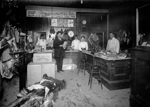 Interiøret til en slaktebutikk. Tre menn og en kvinne står ved disken. En kunde holder en pakke i armen. Skrotter henger på veggen til venstre. På disken til høyre ligger store kjøttstykker på fat. I forgrunn en delvis partert kalv med skinnet på. 
Påskrift bakpå: Lady standing, Maude (Ferguson) Benjamin in Hetherington's Meat Market Hastings. Worked there until she was 20 or 22. Worked there for 6 years.