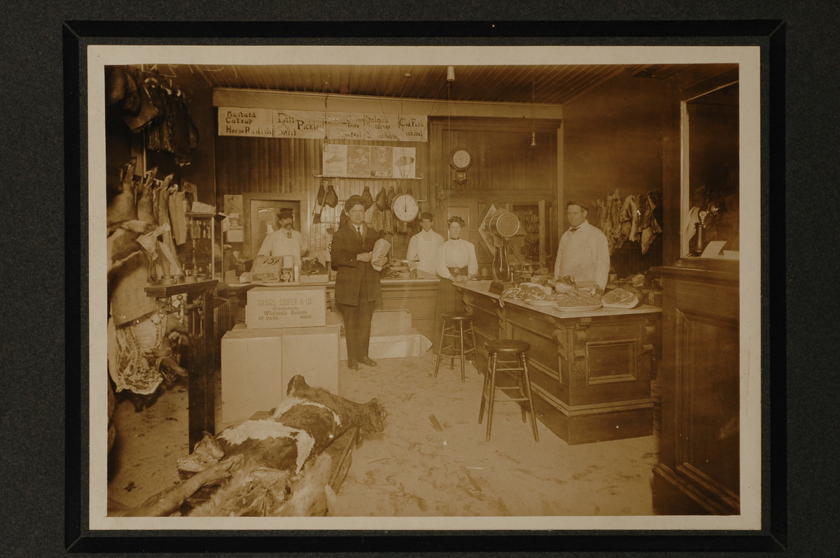 Interiøret til en slaktebutikk. Tre menn og en kvinne står ved disken. En kunde holder en pakke i armen. Skrotter henger på veggen til venstre. På disken til høyre ligger store kjøttstykker på fat. I forgrunn en delvis partert kalv med skinnet på. 
Påskrift bakpå: Lady standing, Maude (Ferguson) Benjamin in Hetherington's Meat Market Hastings. Worked there until she was 20 or 22. Worked there for 6 years. 