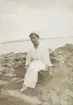 En kvinna i ljus, prickig klänning med volanger, har slagit sig ner på en sten vid en sjökant (Helgasjön?).