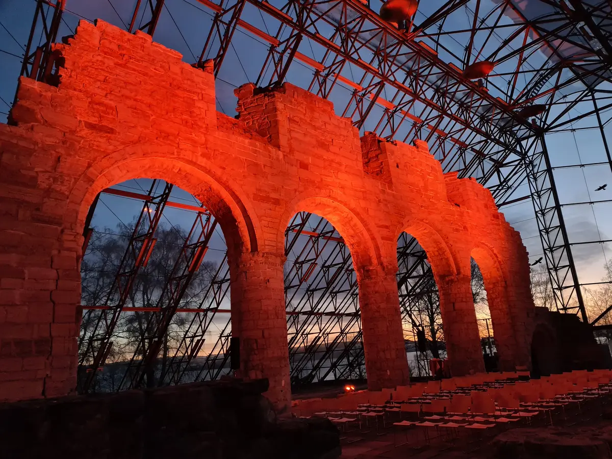 Søylerekka som står igjen etter middelalderkatedralen på Domkirkeodden, er lyssatt med oransje lys. Ble, gråblå kveldshimmel vises gjennom glasset i vernebygget, og det oransje lyset spres også på metallrammene i vernebygget.