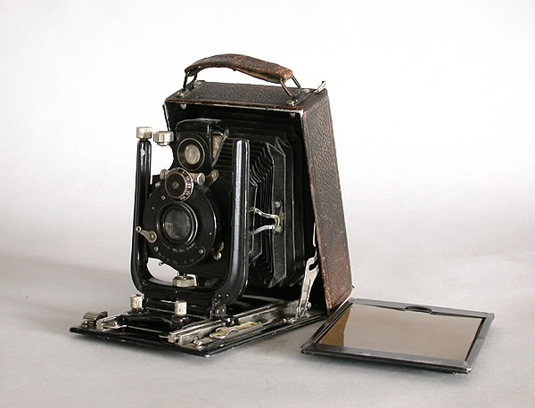 "Kamera-bälg.a) Rektangulär plåtlåda överdragen med svart vaxduk. Ena långsidan fälls utoch bälg med optik dras fram. Optiken kan förskjutas i höjd- och sidoled.Märke: ""Ica"" . Modell: ""Trona 210"".Längd (öppen): 27 cm. Längd (stängd): 15,5 cm.Bredd (öppen): 11 cm.Bredd (stängd): 11 cm.Höjd (öppen): 15,5 cm.Höjd (stängd): 5 cm.b) Löstagbart bakstycke med matt skiva. Kassett för glasplåtar 9x12 cm saknas.Mått: 10x15 cm.c) Trådutlösare, metallsnodd överdragen med svart textil.Längd: 27,5 cm.d) Gulgrön-filter av glas i en metallram som spänns fast runt objektivet.Höjd: 1,5 cm.Diameter: 3,8 cm.e) Oval pung i brunt skinn, försluts med en dragkedja. Rymmer trådutlösarenoch filtret.Bredd: 9 cm.Höjd: 6 cm."