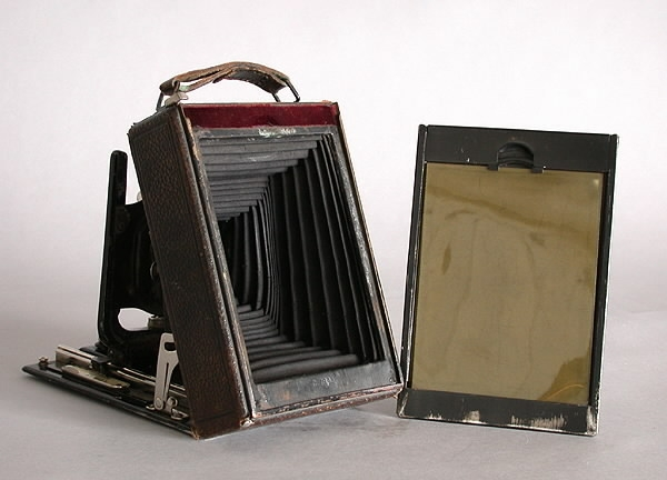 "Kamera-bälg.a) Rektangulär plåtlåda överdragen med svart vaxduk. Ena långsidan fälls utoch bälg med optik dras fram. Optiken kan förskjutas i höjd- och sidoled.Märke: ""Ica"" . Modell: ""Trona 210"".Längd (öppen): 27 cm. Längd (stängd): 15,5 cm.Bredd (öppen): 11 cm.Bredd (stängd): 11 cm.Höjd (öppen): 15,5 cm.Höjd (stängd): 5 cm.b) Löstagbart bakstycke med matt skiva. Kassett för glasplåtar 9x12 cm saknas.Mått: 10x15 cm.c) Trådutlösare, metallsnodd överdragen med svart textil.Längd: 27,5 cm.d) Gulgrön-filter av glas i en metallram som spänns fast runt objektivet.Höjd: 1,5 cm.Diameter: 3,8 cm.e) Oval pung i brunt skinn, försluts med en dragkedja. Rymmer trådutlösarenoch filtret.Bredd: 9 cm.Höjd: 6 cm."