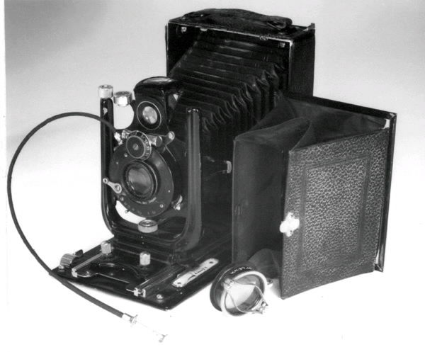 "Kamera-bälg.a) Rektangulär plåtlåda överdragen med svart vaxduk. Ena långsidan fälls utoch bälg med optik dras fram. Optiken kan förskjutas i höjd- och sidoled.Märke: ""Ica"" . Modell: ""Trona 210"".Längd (öppen): 27 cm. Längd (stängd): 15,5 cm.Bredd (öppen): 11 cm.Bredd (stängd): 11 cm.Höjd (öppen): 15,5 cm.Höjd (stängd): 5 cm.b) Löstagbart bakstycke med matt skiva. Kassett för glasplåtar 9x12 cm saknas.Mått: 10x15 cm.c) Trådutlösare, metallsnodd överdragen med svart textil.Längd: 27,5 cm.d) Gulgrön-filter av glas i en metallram som spänns fast runt objektivet.Höjd: 1,5 cm.Diameter: 3,8 cm.e) Oval pung i brunt skinn, försluts med en dragkedja. Rymmer trådutlösarenoch filtret.Bredd: 9 cm.Höjd: 6 cm."