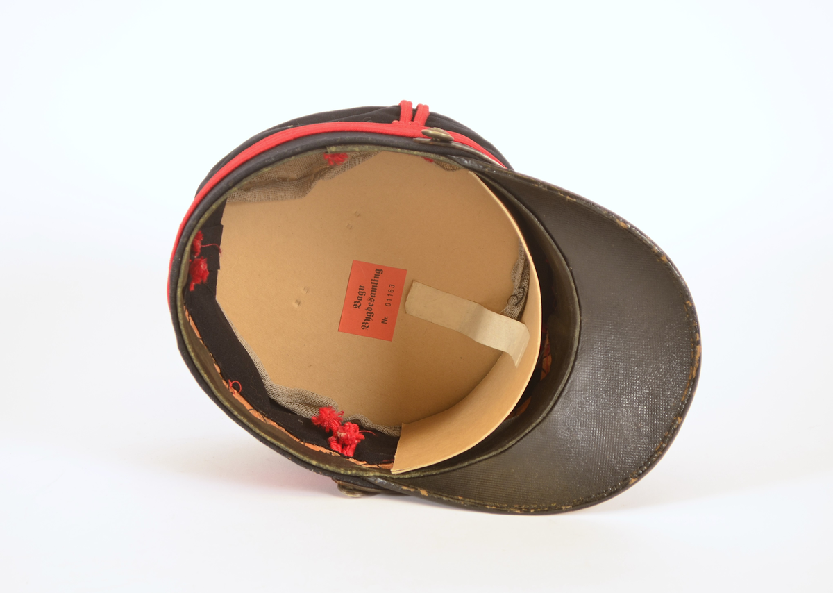 Uniformslue med brem. Over bremmen rem med spenne festet med knapp på hver side. Røde pyntebånd på luen. Knapp pg dusk i front.