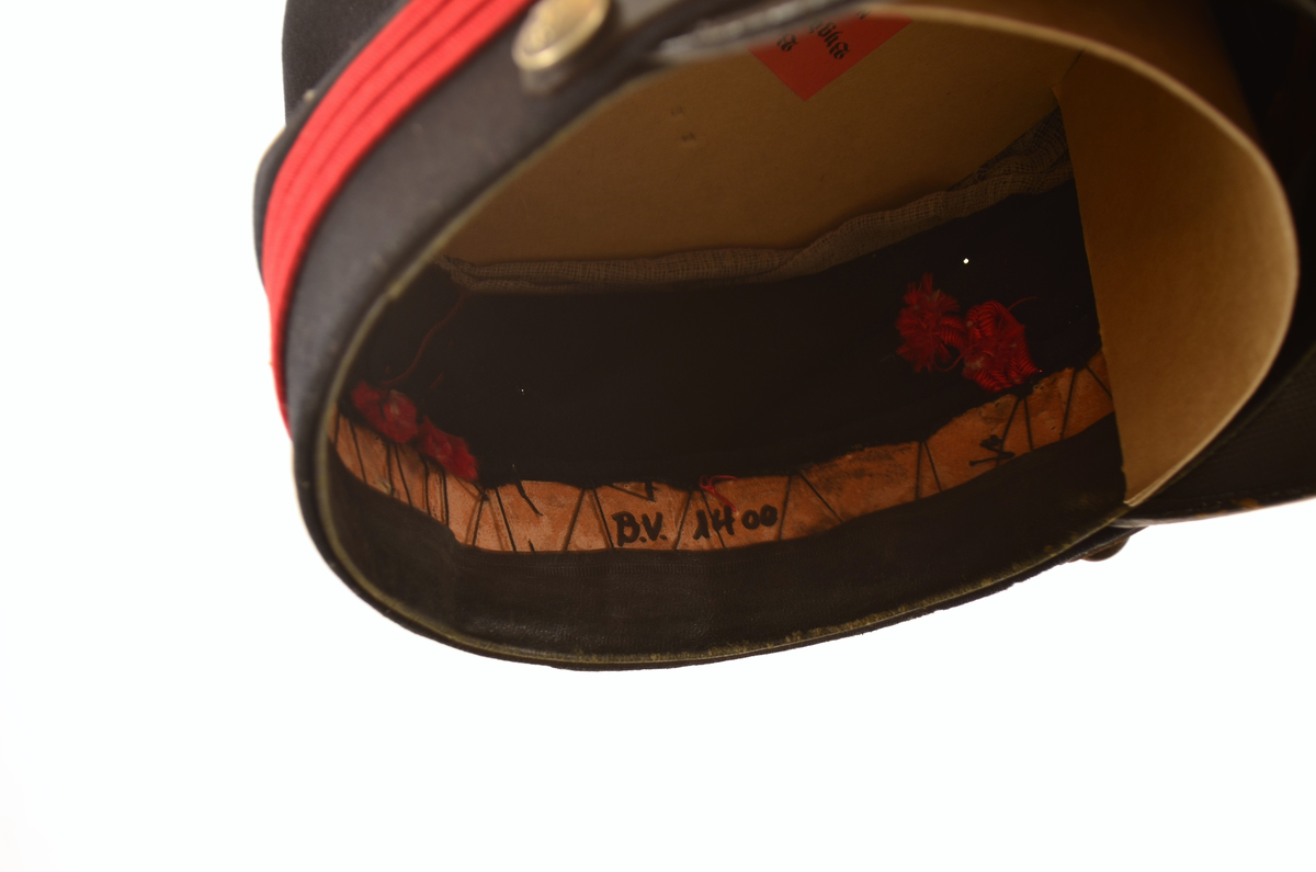 Uniformslue med brem. Over bremmen rem med spenne festet med knapp på hver side. Røde pyntebånd på luen. Knapp pg dusk i front.