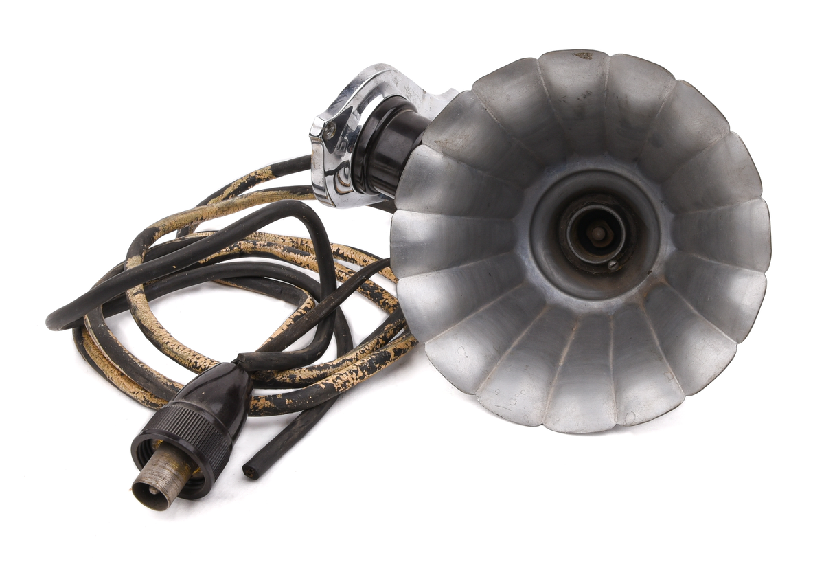 Elektrisk armatur av metall. Troligen modell nr 3050 (SJ typ X), tillverkad av Bröderna Malmströms Metallvarufabrik.
Beskrivning från deras katalog över Järnvägsarmaturer från 1934:
"Fästplattans höjd 160 mm, största bredd 80 mm. Största avstånd från väggen 230 mm. Utföres i gjuten mässing. Skärmen av mässingsplåt, 155 mm diam., invändigt vitlackerad. Lamphållaren är av samma konstruktion som hos n:r 3035, således Edisongänga och fjädrande bottenkontakt. Avsedd för max. 40 volt. Strömbrytaren av typ "Membra". Kan även levereras med normal bajonettlamphållare."