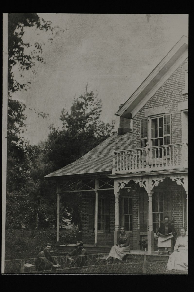 Murhus med to etasjer og forseggjort balkong og veranda. En mann og to kvinner sitter på verandaen. Mannen sitter på en stol og holder avisen. To menn sitter på plenen foran. Nettinggjerde i forkant. 