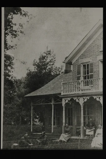 Murhus med to etasjer og forseggjort balkong og veranda. En mann og to kvinner sitter på verandaen. Mannen sitter på en stol og holder avisen. To menn sitter på plenen foran. Nettinggjerde i forkant.