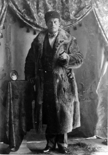 Portrett av en mann med bart og pelsfrakk som holder en sigar. Han lener høyre hånd på et lite bord med ovalt bilde av et barn på. Drapert gardin i bakgrunn.