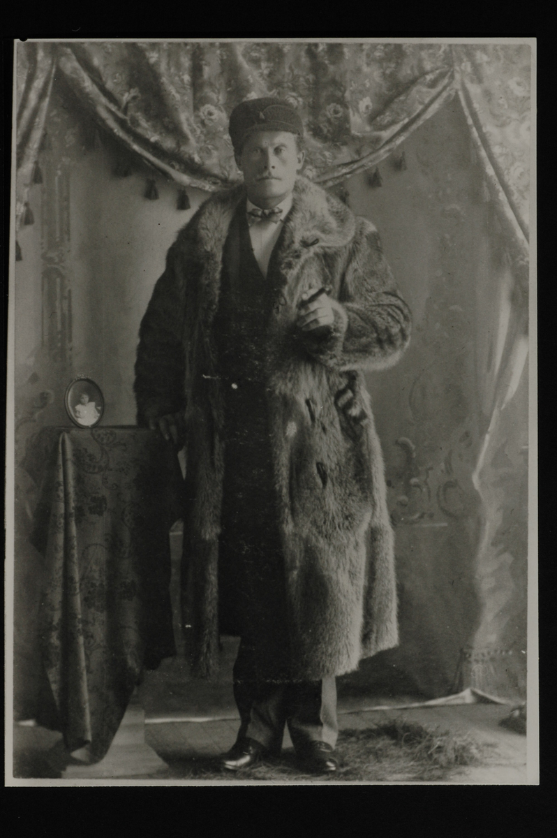 Portrett av en mann med bart og pelsfrakk som holder en sigar. Han lener høyre hånd på et lite bord med ovalt bilde av et barn på. Drapert gardin i bakgrunn. 