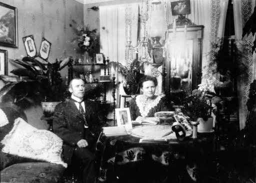 Interiør med mann og kvinne som sitter ved et rundt bord. På bordet bøker, bilder og en stereograf. En sofa med en pute til venstre. Flere planter på pidestaller. Bilder og et speil på veggen. En majolika-lampe henger over bordet. Lyse gardinger foran vinduene. Påskrift bakpå: Stua til taxi-sjåfør i Egersund.