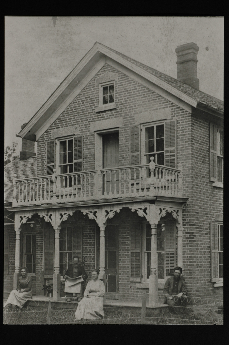 Murhus i to etasjer med forseggjort veranda. To kvinner og to menn sitter på verandaen. En mann holder en avis. Nettinggjerde i forkant.  