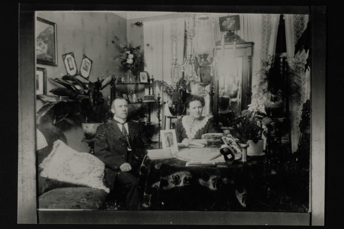 Interiør med mann og kvinne som sitter ved et rundt bord. På bordet bøker, bilder og en stereograf. En sofa med en pute til venstre. Flere planter på pidestaller. Bilder og et speil på veggen. En majolika-lampe henger over bordet. Lyse gardinger foran vinduene. Påskrift bakpå: Stua til taxi-sjåfør i Egersund.