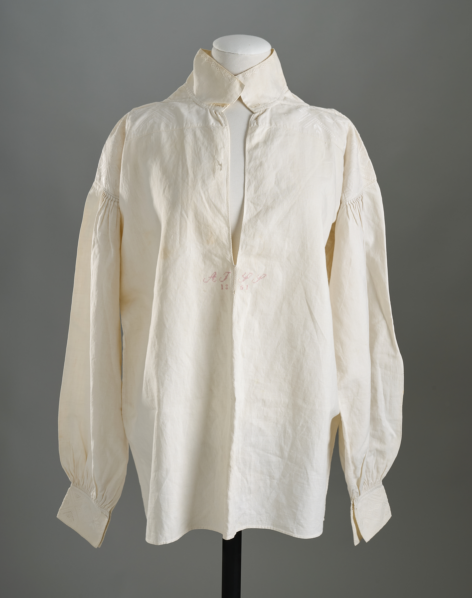 Linskjorte med rettstående krave og hvitsømsbroderi på bærestykke, krave og mansjetter. Innfelte trekanter på bærestykket over skuldre mot kraven. Kraven har to knapphull og er sammenrynket MF mot halsåpning. Splitt MF med pyntesøm nederst. Initialer i rosa garn med dronningsting, årstall med korssting under splitten. Ermene er isydde med 11 legg foran og bak. Isydde spjeld under ermhulene. Ermene er tett rynket til mansjettene som også har to knapphull i åpningen. Taggekant på krave og mansjetter. Smal fald i nedre kant.