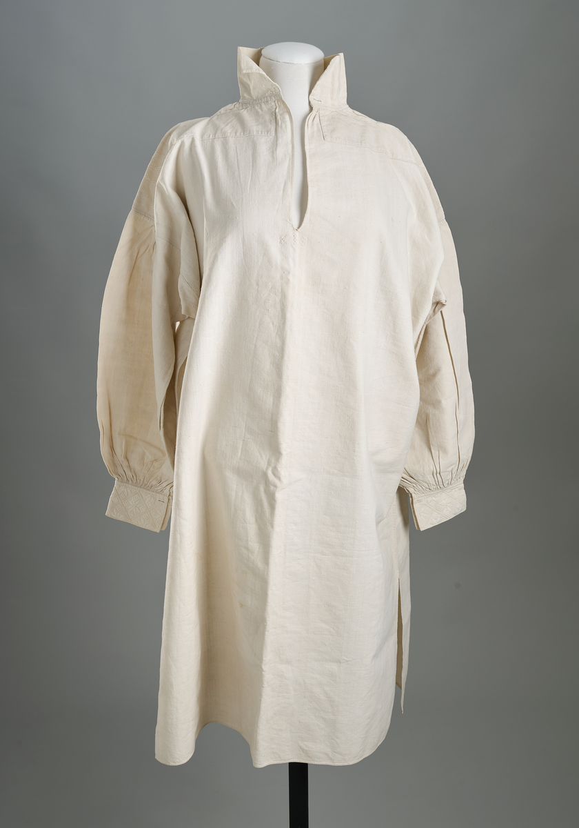 Linskjorte med rettstående krave og hvitsømsbroderi på bærestykke og mansjetter. Innfelte trekanter på bærestykket over skuldre mot kraven. Kraven har to knapphull mot halsåpning. Splitt i begge sider. Initialer i hvitt garn med korssting under splitten. Ermene er isydde med 3 legg foran og bak. Isydde spjeld under ermhulene. Ermene er tett rynket til mansjettene som også har to knapphull ved åpningen. Taggekant på mansjetter. Smal fald i nedre kant.