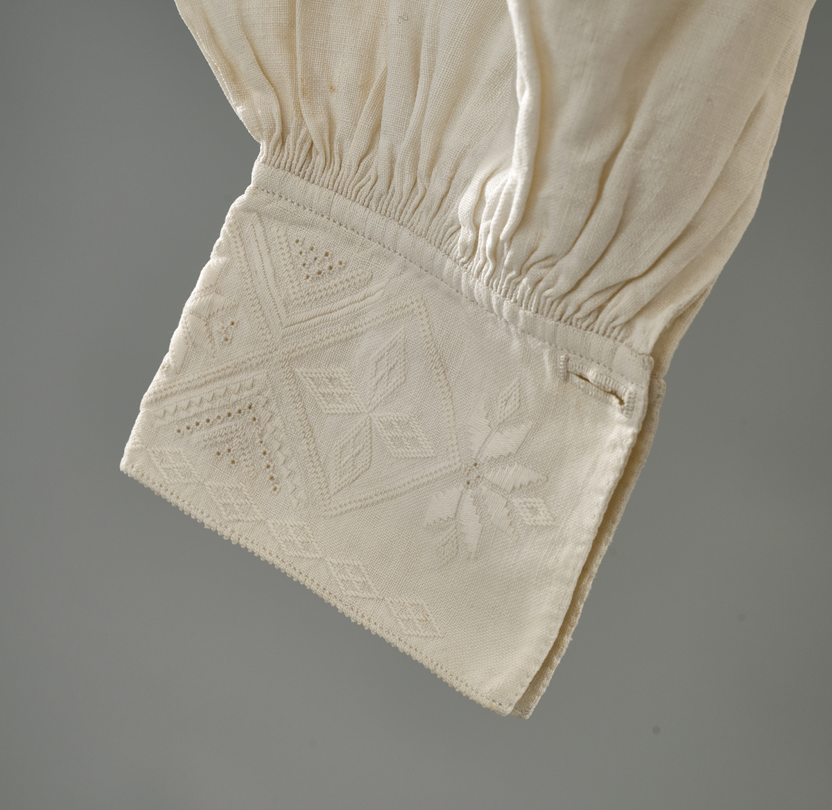 Linskjorte med rettstående krave og hvitsømsbroderi på bærestykke, krave og mansjetter. Innfelte trekanter på bærestykket over skuldre mot kraven. Kraven har to knapphull og er sammenrynket MF mot halsåpning. Splitt MF med pyntesøm nederst. Initialer i rosa garn med dronningsting, årstall med korssting under splitten. Ermene er isydde med 11 legg foran og bak. Isydde spjeld under ermhulene. Ermene er tett rynket til mansjettene som også har to knapphull i åpningen. Taggekant på krave og mansjetter. Smal fald i nedre kant.