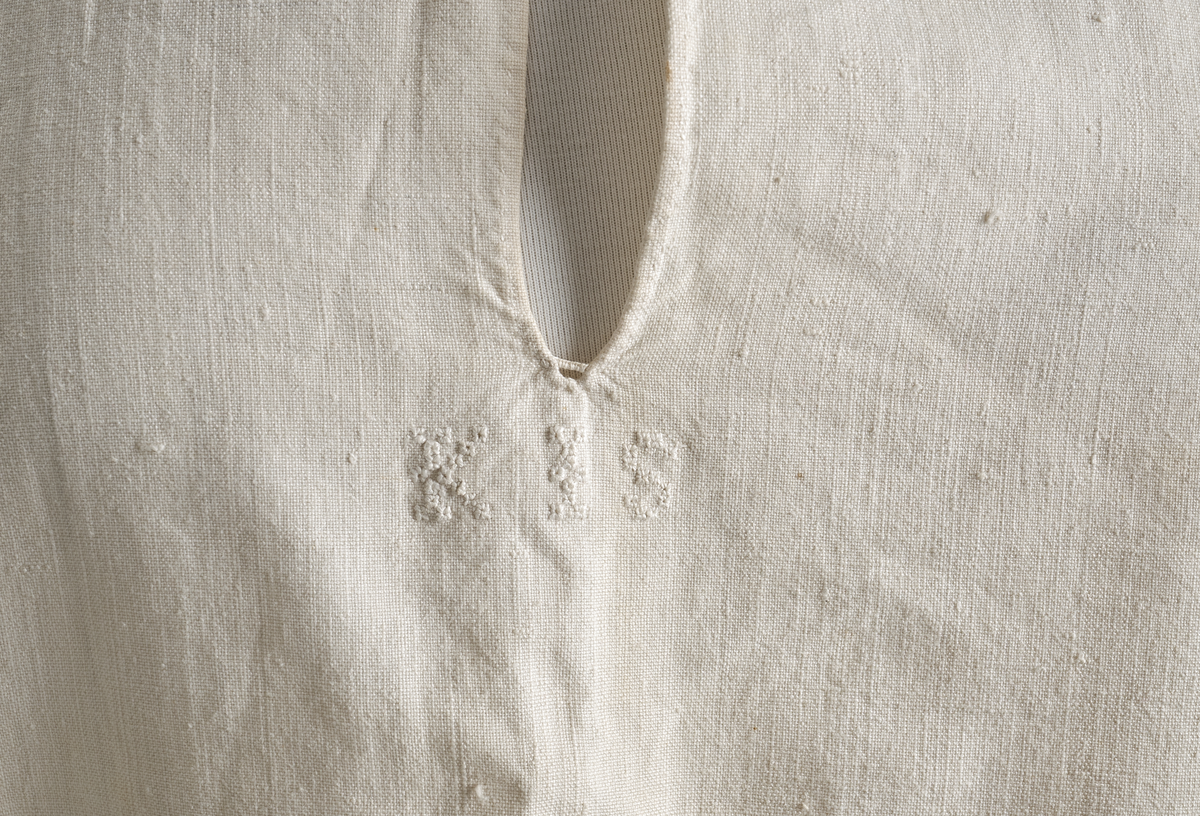 Linskjorte med rettstående krave og hvitsømsbroderi på bærestykke og mansjetter. Innfelte trekanter på bærestykket over skuldre mot kraven. Kraven har to knapphull mot halsåpning. Splitt i begge sider. Initialer i hvitt garn med korssting under splitten. Ermene er isydde med 3 legg foran og bak. Isydde spjeld under ermhulene. Ermene er tett rynket til mansjettene som også har to knapphull ved åpningen. Taggekant på mansjetter. Smal fald i nedre kant.