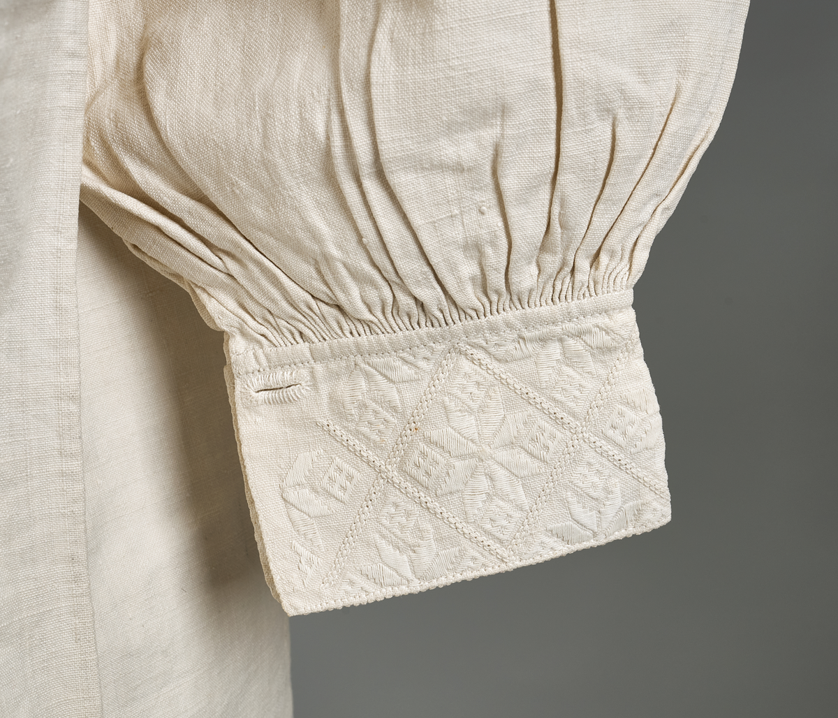 Linskjorte med rettstående krave og hvitsømsbroderi på bærestykke og mansjetter. Innfelte trekanter på bærestykket over skuldre mot kraven. Kraven har to knapphull mot halsåpning. Splitt i begge sider. Initialer i hvitt garn med korssting under splitten. Ermene er isydde med 3 legg foran og bak. Isydde spjeld under ermhulene. Ermene er tett rynket til mansjettene som også har to knapphull ved åpningen. Taggekant på mansjetter. Smal fald i nedre kant.