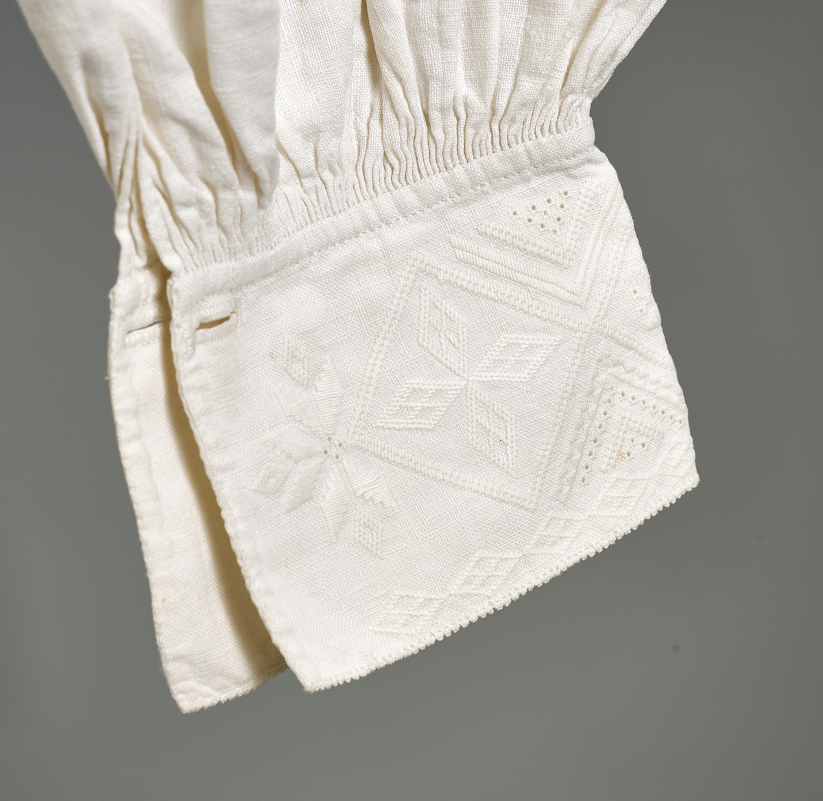 Linskjorte med rettstående krave og hvitsømsbroderi på bærestykke, krave og mansjetter. Innfelte trekanter på bærestykket over skuldre mot kraven. Kraven har to knapphull og er sammenrynket MF mot halsåpning. Splitt MF med pyntesøm nederst. Initialer i rosa garn med dronningsting, årstall med korssting under splitten. Ermene er isydde med 11 legg foran og bak. Isydde spjeld under ermhulene. Ermene er tett rynket til mansjettene som også har to knapphull i åpningen. Taggekant på krave og mansjetter. Smal fald i nedre kant.