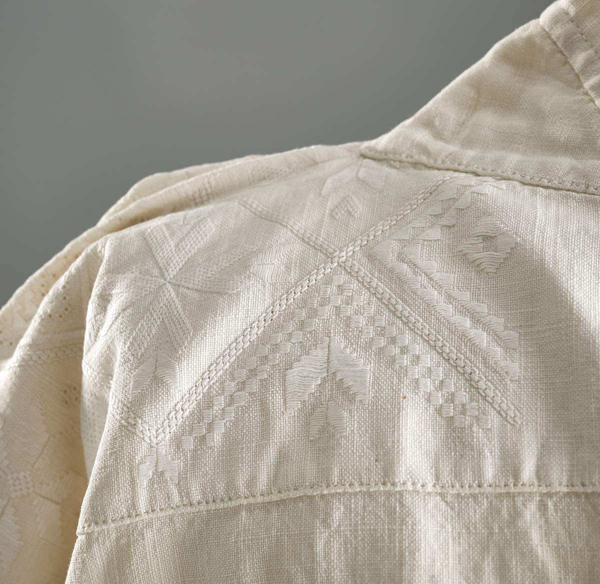 Linskjorte med rettstående krave og hvitsømsbroderi på bærestykke, krave og mansjetter. Innfelte trekanter på bærestykket over skuldre mot kraven. Kraven har to knapphull og er sammenrynket MF mot halsåpning. Splitt MF med pyntesøm nederst. Initialer i rosa garn med dronningsting, årstall med korssting under splitten. Ermene er isydde med 11 legg foran og bak. Isydde spjeld under ermhulene. Ermene er tett rynket til mansjettene som også har to knapphull i åpningen. Taggekant på krave og mansjetter. Smal fald i nedre kant.