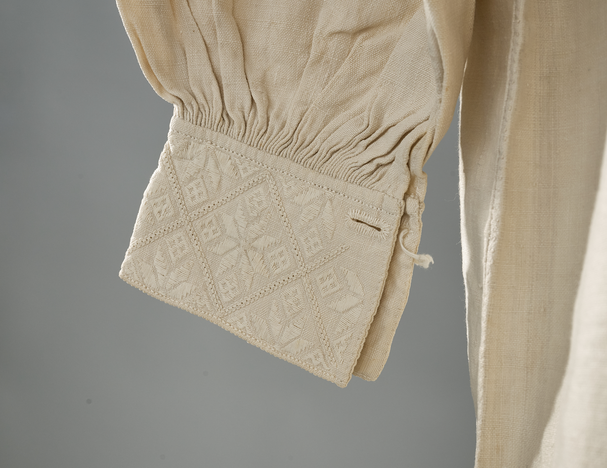 Linskjorte med rettstående krave og hvitsømsbroderi på bærestykke og mansjetter. Innfelte trekanter på bærestykket over skuldre mot kraven. Kraven har to knapphull mot halsåpning. Splitt i begge sider. Initialer i hvitt garn med korssting under splitten. Ermene er isydde med 3 legg foran og bak. Isydde spjeld under ermhulene. Ermene er tett rynket til mansjettene som også har to knapphull ved åpningen. Taggekant på mansjetter. Smal fald i nedre kant.