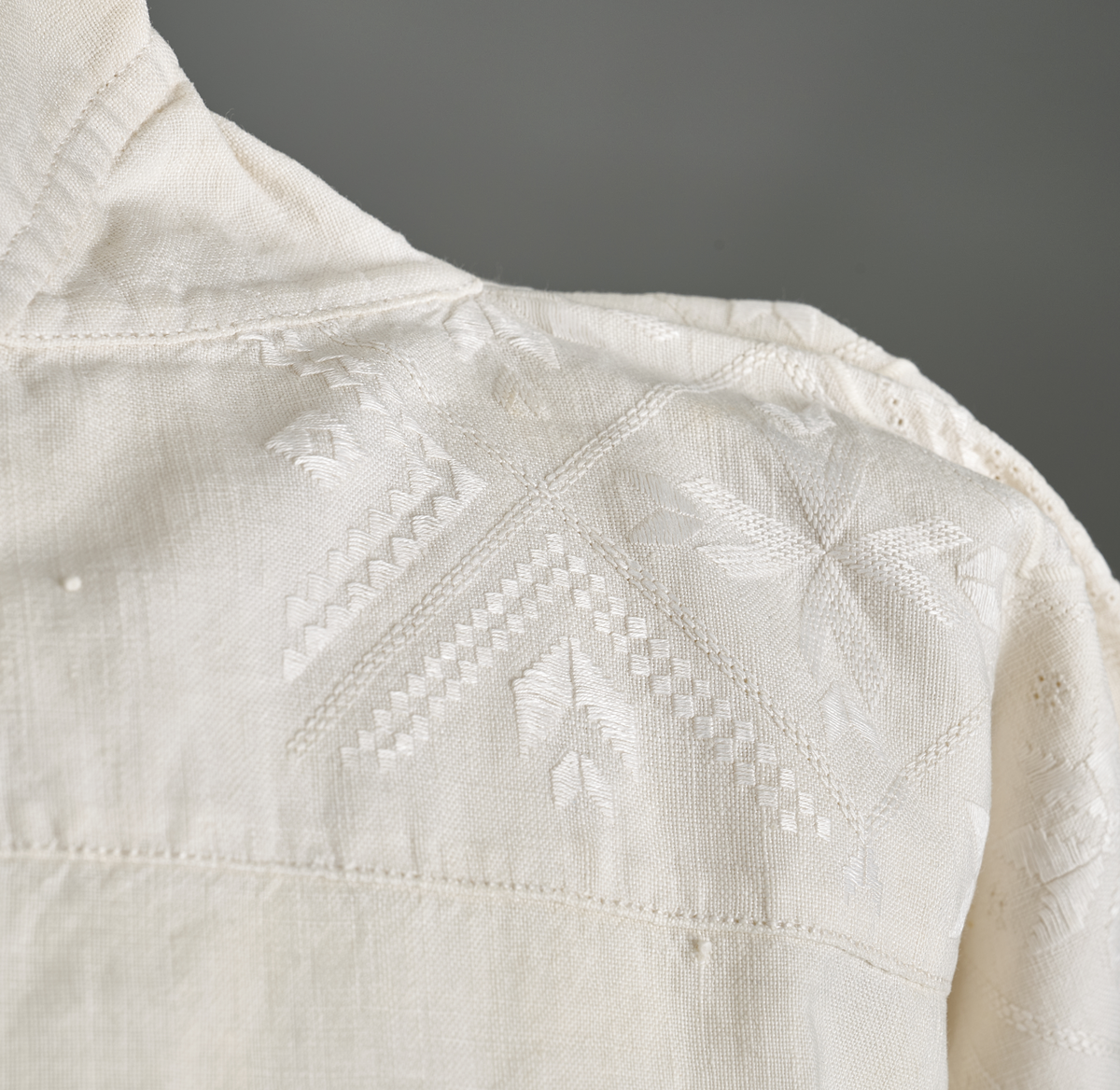 Linskjorte med rettstående krave og hvitsømsbroderi på bærestykke, krave og mansjetter. Innfelte trekanter på bærestykket over skuldre mot kraven. Kraven har to knapphull og er sammenrynket MF mot halsåpning. Splitt MF med pyntesøm nederst. Initialer i rosa garn med dronningsting, årstall med korssting under splitten. Ermene er isydde med 11 legg foran og bak. Isydde spjeld under ermhulene. Ermene er tett rynket til mansjettene som også har to knapphull i åpningen. Taggekant på krave og mansjetter. Smal fald i nedre kant.