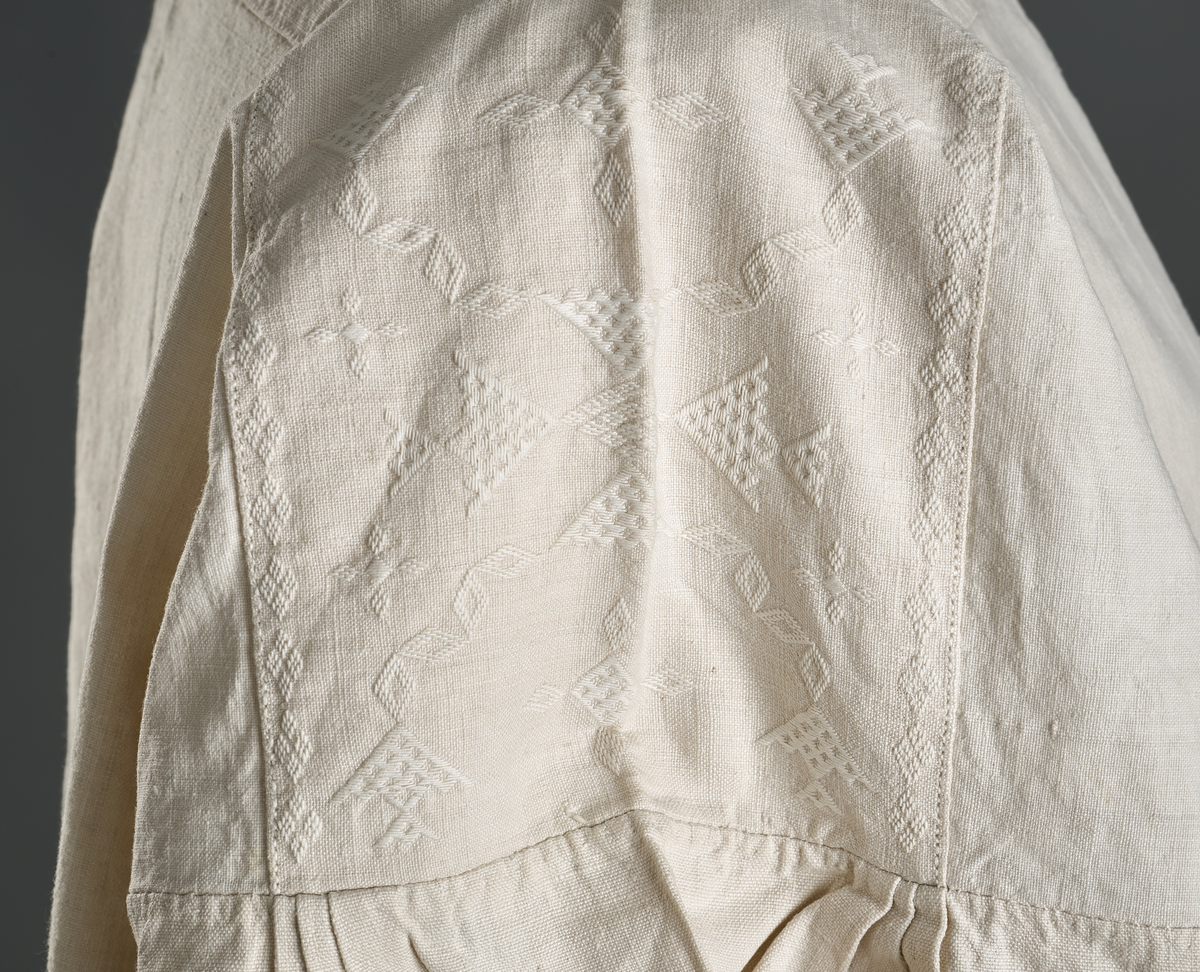 Linskjorte med rettstående krave og hvitsømsbroderi på bærestykke og mansjetter. Innfelte trekanter på bærestykket over skuldre mot kraven. Kraven har to knapphull mot halsåpning. Splitt i begge sider. Initialer i hvitt garn med korssting under splitten. Ermene er isydde med 3 legg foran og bak. Isydde spjeld under ermhulene. Ermene er tett rynket til mansjettene som også har to knapphull ved åpningen. Taggekant på mansjetter. Smal fald i nedre kant.