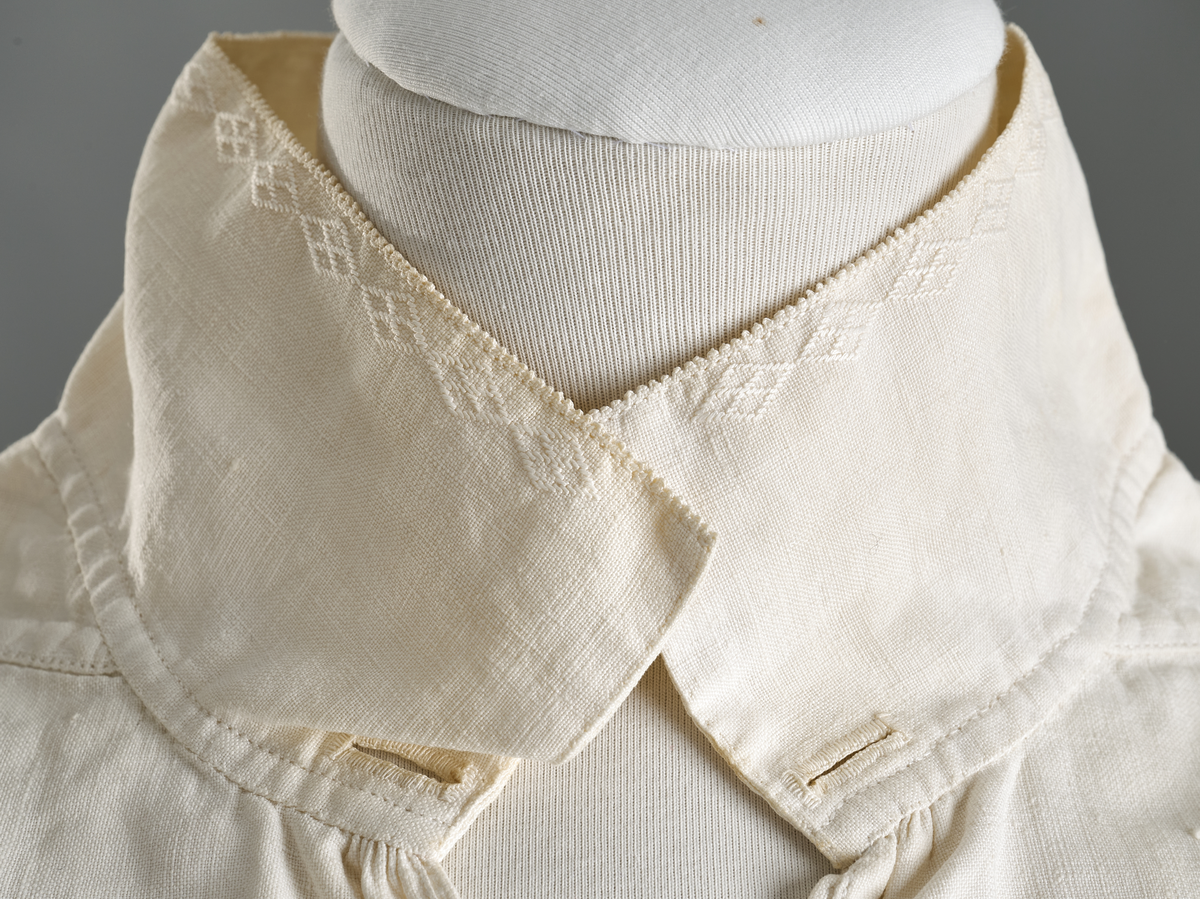 Linskjorte med rettstående krave og hvitsømsbroderi på bærestykke, krave og mansjetter. Innfelte trekanter på bærestykket over skuldre mot kraven. Kraven har to knapphull og er sammenrynket MF mot halsåpning. Splitt MF med pyntesøm nederst. Initialer i rosa garn med dronningsting, årstall med korssting under splitten. Ermene er isydde med 11 legg foran og bak. Isydde spjeld under ermhulene. Ermene er tett rynket til mansjettene som også har to knapphull i åpningen. Taggekant på krave og mansjetter. Smal fald i nedre kant.