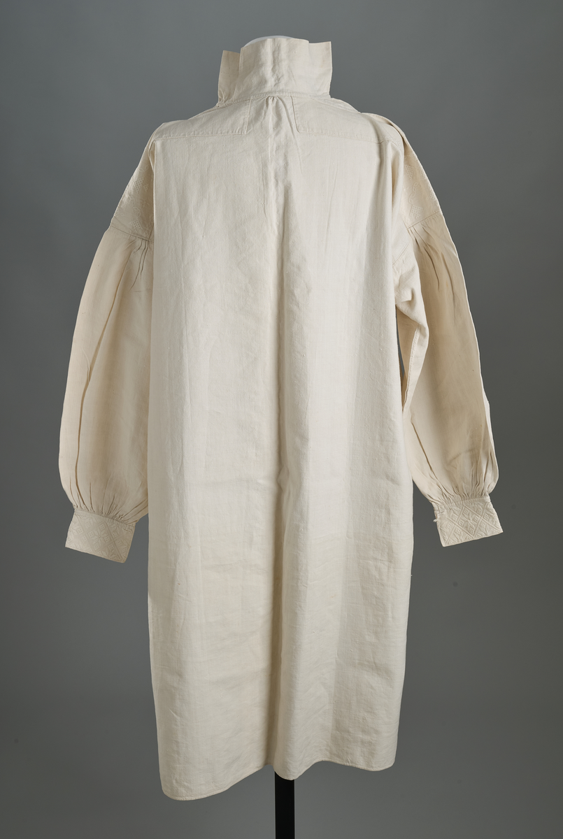 Linskjorte med rettstående krave og hvitsømsbroderi på bærestykke og mansjetter. Innfelte trekanter på bærestykket over skuldre mot kraven. Kraven har to knapphull mot halsåpning. Splitt i begge sider. Initialer i hvitt garn med korssting under splitten. Ermene er isydde med 3 legg foran og bak. Isydde spjeld under ermhulene. Ermene er tett rynket til mansjettene som også har to knapphull ved åpningen. Taggekant på mansjetter. Smal fald i nedre kant.