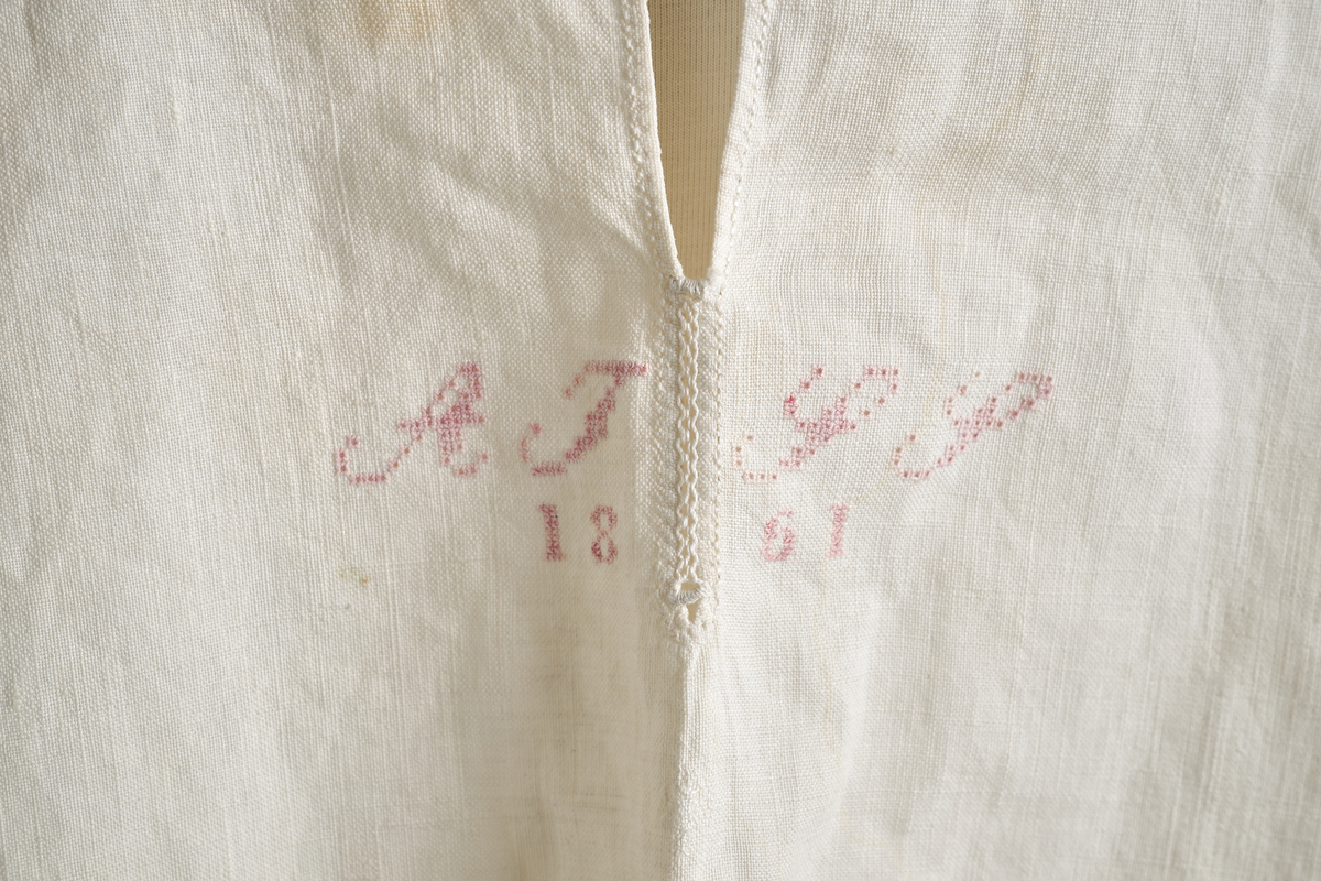 Linskjorte med rettstående krave og hvitsømsbroderi på bærestykke, krave og mansjetter. Innfelte trekanter på bærestykket over skuldre mot kraven. Kraven har to knapphull og er sammenrynket MF mot halsåpning. Splitt MF med pyntesøm nederst. Initialer i rosa garn med dronningsting, årstall med korssting under splitten. Ermene er isydde med 11 legg foran og bak. Isydde spjeld under ermhulene. Ermene er tett rynket til mansjettene som også har to knapphull i åpningen. Taggekant på krave og mansjetter. Smal fald i nedre kant.