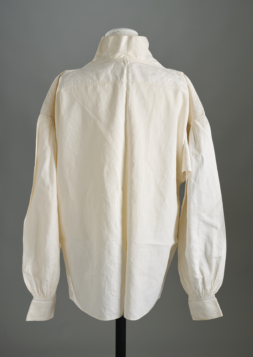 Linskjorte med rettstående krave og hvitsømsbroderi på bærestykke, krave og mansjetter. Innfelte trekanter på bærestykket over skuldre mot kraven. Kraven har to knapphull og er sammenrynket MF mot halsåpning. Splitt MF med pyntesøm nederst. Initialer i rosa garn med dronningsting, årstall med korssting under splitten. Ermene er isydde med 11 legg foran og bak. Isydde spjeld under ermhulene. Ermene er tett rynket til mansjettene som også har to knapphull i åpningen. Taggekant på krave og mansjetter. Smal fald i nedre kant.