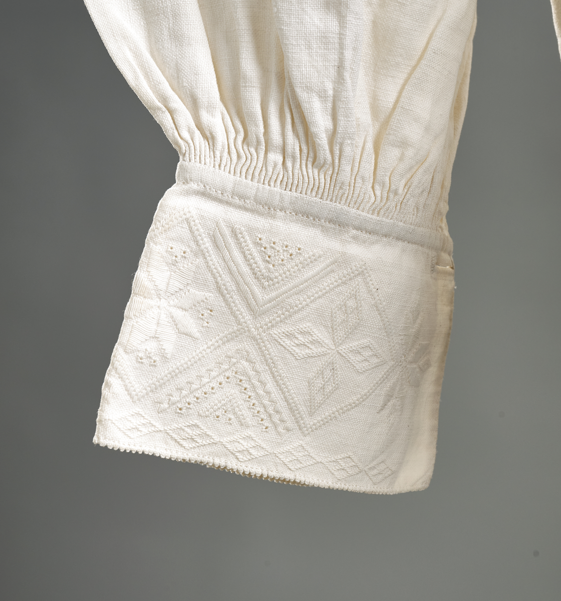 Linskjorte med rettstående krave og hvitsømsbroderi på bærestykke, krave og mansjetter. Innfelte trekanter på bærestykket over skuldre mot kraven. Kraven har to knapphull og er sammenrynket MF mot halsåpning. Splitt MF med pyntesøm nederst. Initialer i rosa garn med dronningsting, årstall med korssting under splitten. Ermene er isydde med 11 legg foran og bak. Isydde spjeld under ermhulene. Ermene er tett rynket til mansjettene som også har to knapphull i åpningen. Taggekant på krave og mansjetter. Smal fald i nedre kant.