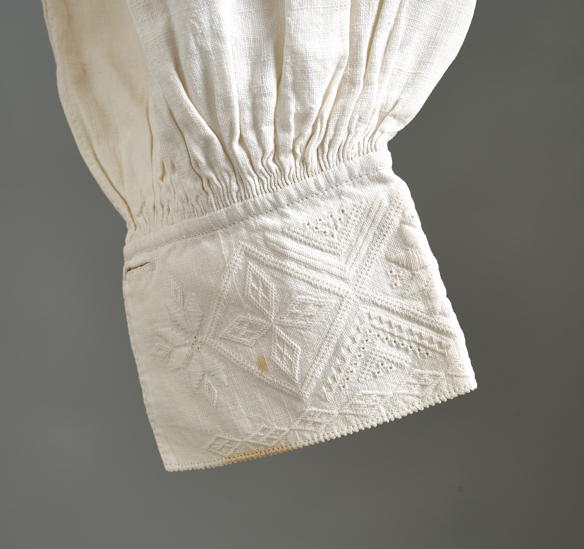 Linskjorte med rettstående krave og hvitsømsbroderi på bærestykke, krave og mansjetter. Innfelte trekanter på bærestykket over skuldre mot kraven. Kraven har to knapphull og er sammenrynket MF mot halsåpning. Splitt MF med pyntesøm nederst. Initialer i rosa garn med dronningsting, årstall med korssting under splitten. Ermene er isydde med 11 legg foran og bak. Isydde spjeld under ermhulene. Ermene er tett rynket til mansjettene som også har to knapphull i åpningen. Taggekant på krave og mansjetter. Smal fald i nedre kant.