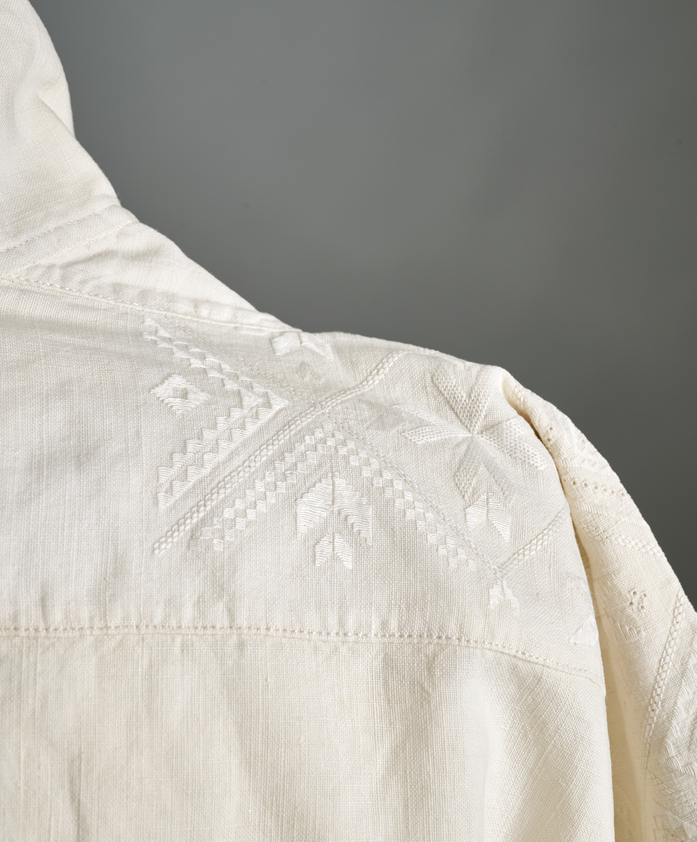 Linskjorte med rettstående krave og hvitsømsbroderi på bærestykke, krave og mansjetter. Innfelte trekanter på bærestykket over skuldre mot kraven. Kraven har to knapphull og er sammenrynket MF mot halsåpning. Splitt MF med pyntesøm nederst. Initialer i rosa garn med dronningsting, årstall med korssting under splitten. Ermene er isydde med 11 legg foran og bak. Isydde spjeld under ermhulene. Ermene er tett rynket til mansjettene som også har to knapphull i åpningen. Taggekant på krave og mansjetter. Smal fald i nedre kant.