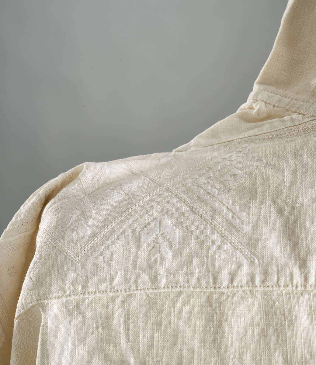 Linskjorte med rettstående krave og hvitsømsbroderi på bærestykke, krave og mansjetter. Innfelte trekanter på bærestykket over skuldre mot kraven. Kraven har to knapphull og er sammenrynket MF mot halsåpning. Splitt MF med pyntesøm nederst. Initialer i rosa garn med dronningsting, årstall med korssting under splitten. Ermene er isydde med 11 legg foran og bak. Isydde spjeld under ermhulene. Ermene er tett rynket til mansjettene som også har to knapphull i åpningen. Taggekant på krave og mansjetter. Smal fald i nedre kant.