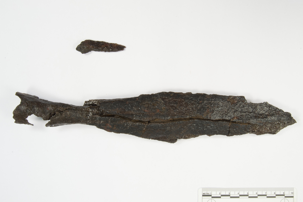 "Spydspiss av jern av typen Jan Petersen l. c., fig. 7 (type A), avbrukket i odden, nuværende lengde 28,5 cm., bladets bredde 5,3 cm." Ny beskrivelse etter revisjon: Spydspiss av jern, Solbergs type VI. 1. Spydspissen er korrodert og oppsprukket, og deler av bladets odd, egger og nedre del er brukket av og mangler, og gjennom falen går et korrosjonshull. To mindre fragmenter foreligger løst. Spyd stl: 290 mm, blad stbr: 53 mm, stt: 22 mm, falens ende diam: 30 mm.