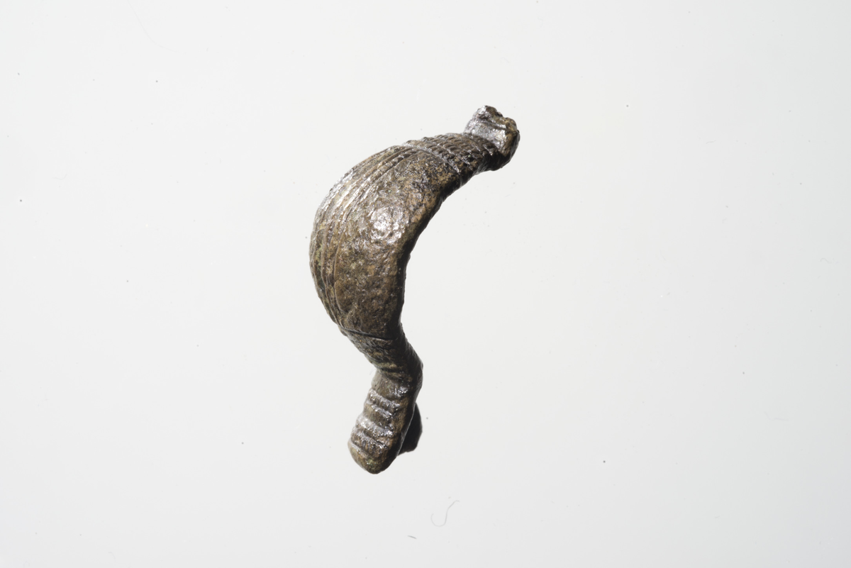 Spenne, likearmet, en kort avrundet endeplate med bevart nålefeste på baksiden. Motstående endeplate er avknekt. Sterkt hvelvet bøyle med fire tverrstilte riflede streker i hver ende ved overgangen til endeplatene med fire i den ene og fem i den andre. Langsgående over midtpartiet går det en sentrert fordypning med én glatt og én riflet strek på hver side. Disse støter mot de tverrgående, riflede strekene på endene og danner to langsgående flater på hver side. Av dekoren på de langsgående flatene er det kun bevart tre fordypninger som ligger langs strekene. På den bevarte endeplaten ses også tverrgående streker: en sentrert fordypning, en glatt og en riflet strek på hver side av denne.