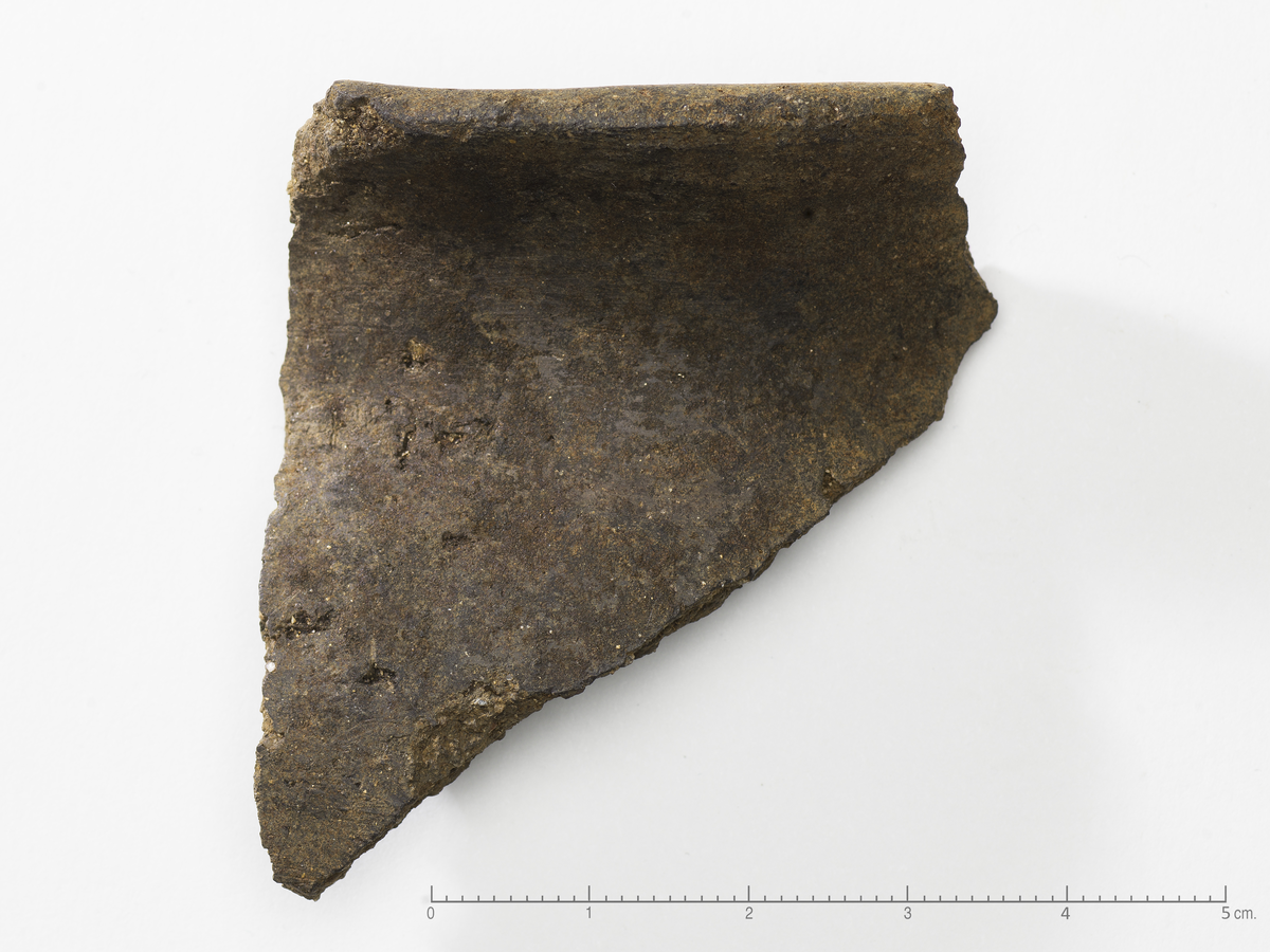 1 Skår fra finere bordkar av fin sandmagret leire. Randskår. Udekorert, men glattpolert. Karet har kort hals, og delvis avflatet rand. Skårets profil er S-formet. Trolig relatert drikkekultur, men vanskelig å typebestemme karet kun ut fra dette ene skåret. Mål: Tykkelse: 0,5 cm. Lengde: 5,7 cm. Bredde: 4 cm.
