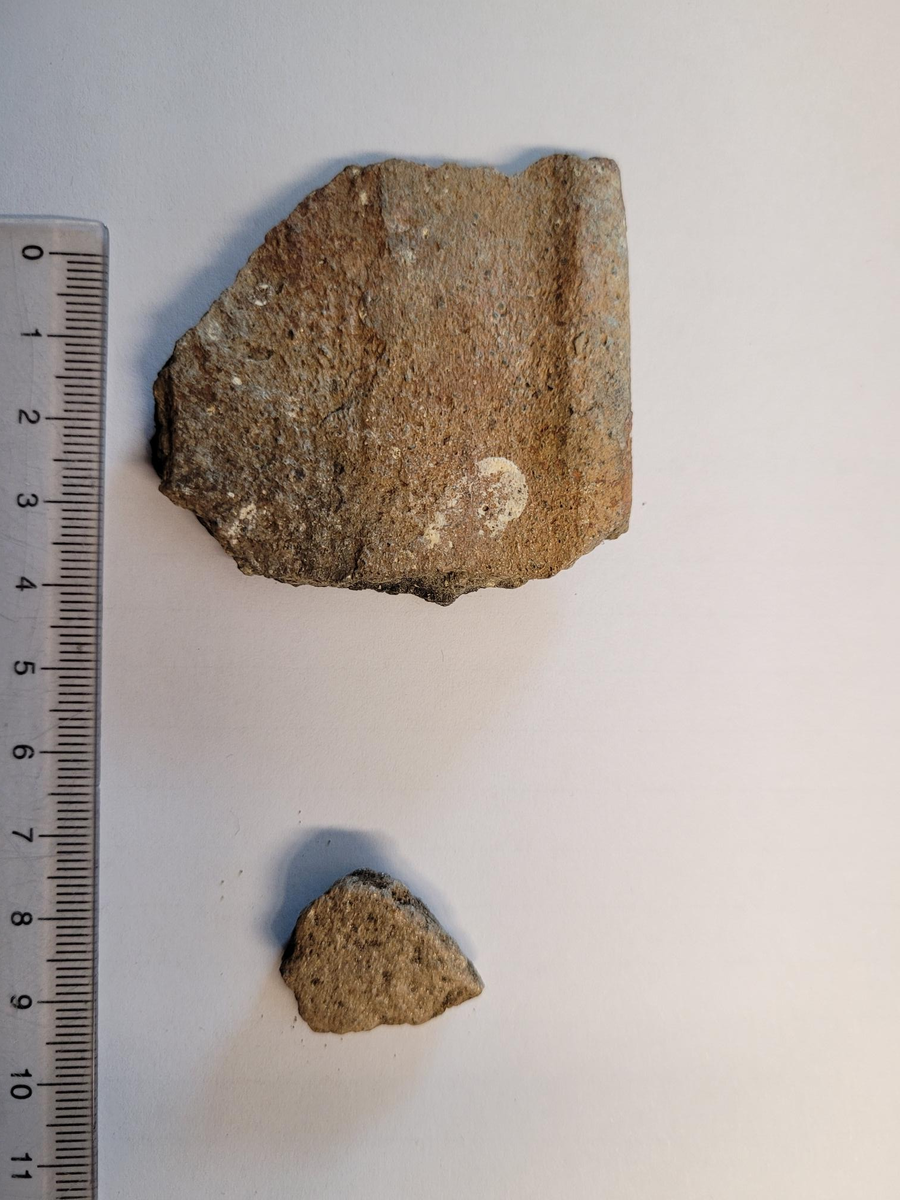 Ett skår av spannformet leirkar. Randskår. Klebermagret. Med rand og øvre del av vegg. Randform: trolig type F (Kristoffersen&Magnus: 2010, fig. 5). Klare tegn på sot/varme på innsida. Utsida har et relativt bredt horisontalfelt/bånd (max. 2,5 cm) under randa, og det finst mulige spor (rødfarging, korrosjon) etter jernbånd her. Også mulige spor etter korrosjon øverst på innsida. Avskalling på nedre bevarte del av utsida, og dermed ingen bevarte spor etter dekor.
Fnr. 200098
