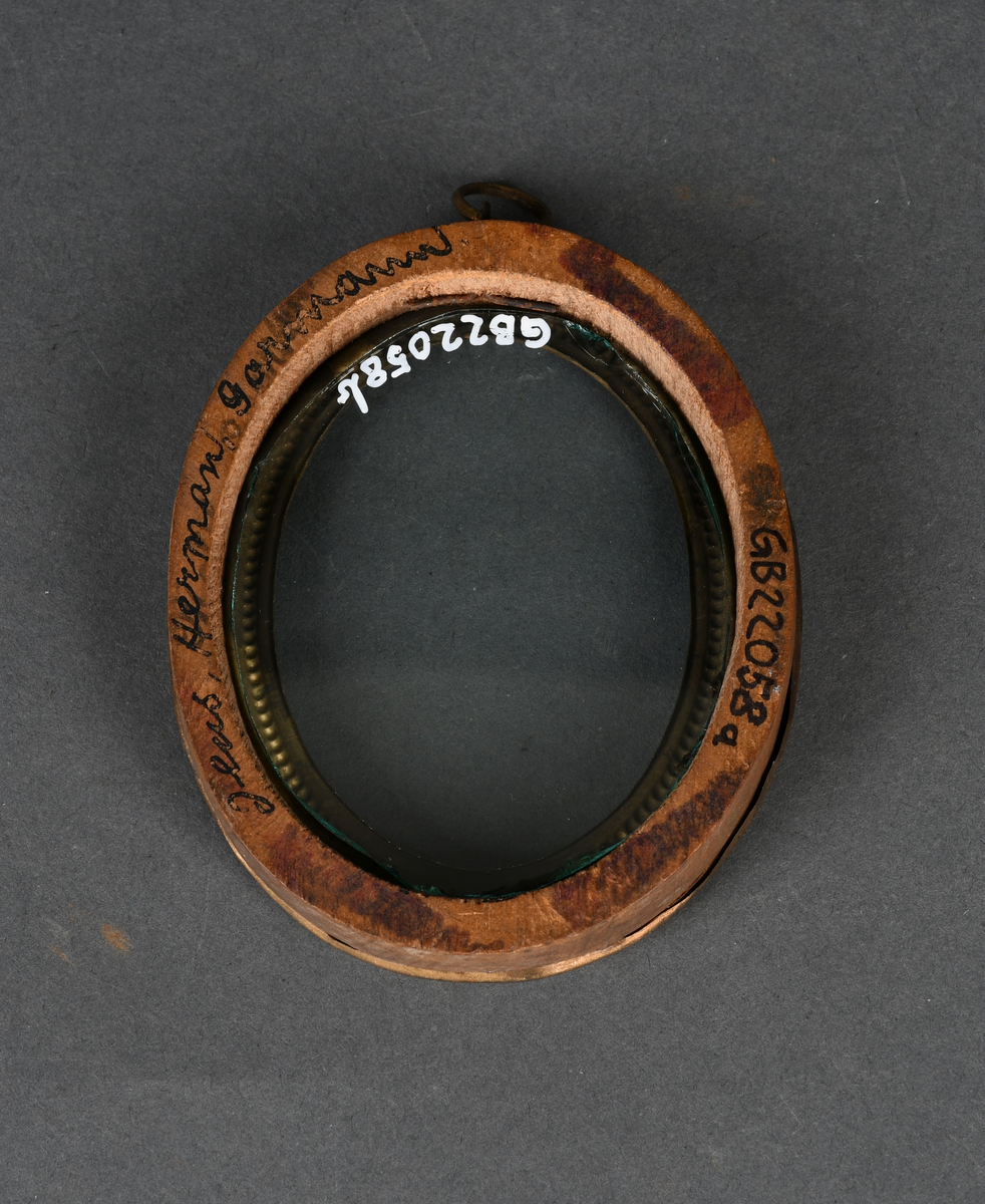 Oval bilderamme med treramme og gullmalt, profilert metallkant med to perlerader. Rammen har et løst, beskyttende glass og en ring festet til øvre kant.