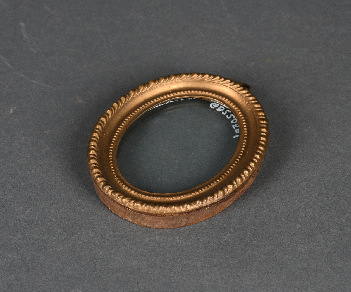 Oval bilderamme med treramme og gullmalt, profilert metallkant med to perlerader. Rammen har et løst, beskyttende glass og en ring festet til øvre kant.