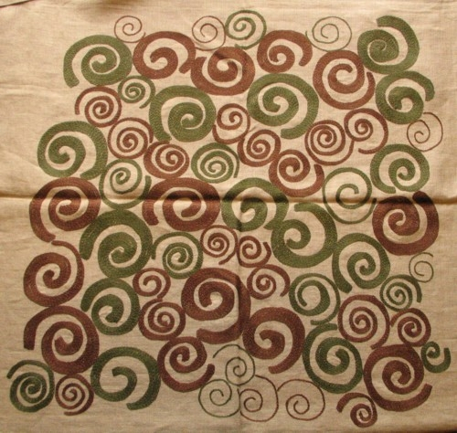 Fyrkantig handbroderad duk, sydd på oblekt grovt linnetyg. Broderier i hörnen samt i mitten (ca 62 x 62 cm) i mörkt grönt och brunt lingarn. Broderierna består av mer och mindre fyllda spiralformer tätt samlade.