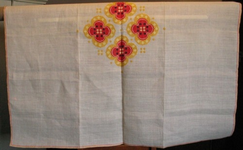 Broderad kvadratisk stor duk. Korsstygn på halvblekt linne med lingarn i rött, gult, svart, ljusgult och vitt.
 Mönstret: Fyra stora vallmoblommor med små prickar emellan placerade på dukens mitt. En röd och en gul rand längs fållarna.