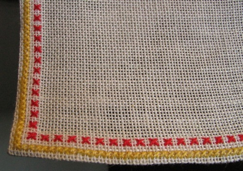 Broderad kvadratisk stor duk. Korsstygn på halvblekt linne med lingarn i rött, gult, svart, ljusgult och vitt.
 Mönstret: Fyra stora vallmoblommor med små prickar emellan placerade på dukens mitt. En röd och en gul rand längs fållarna.